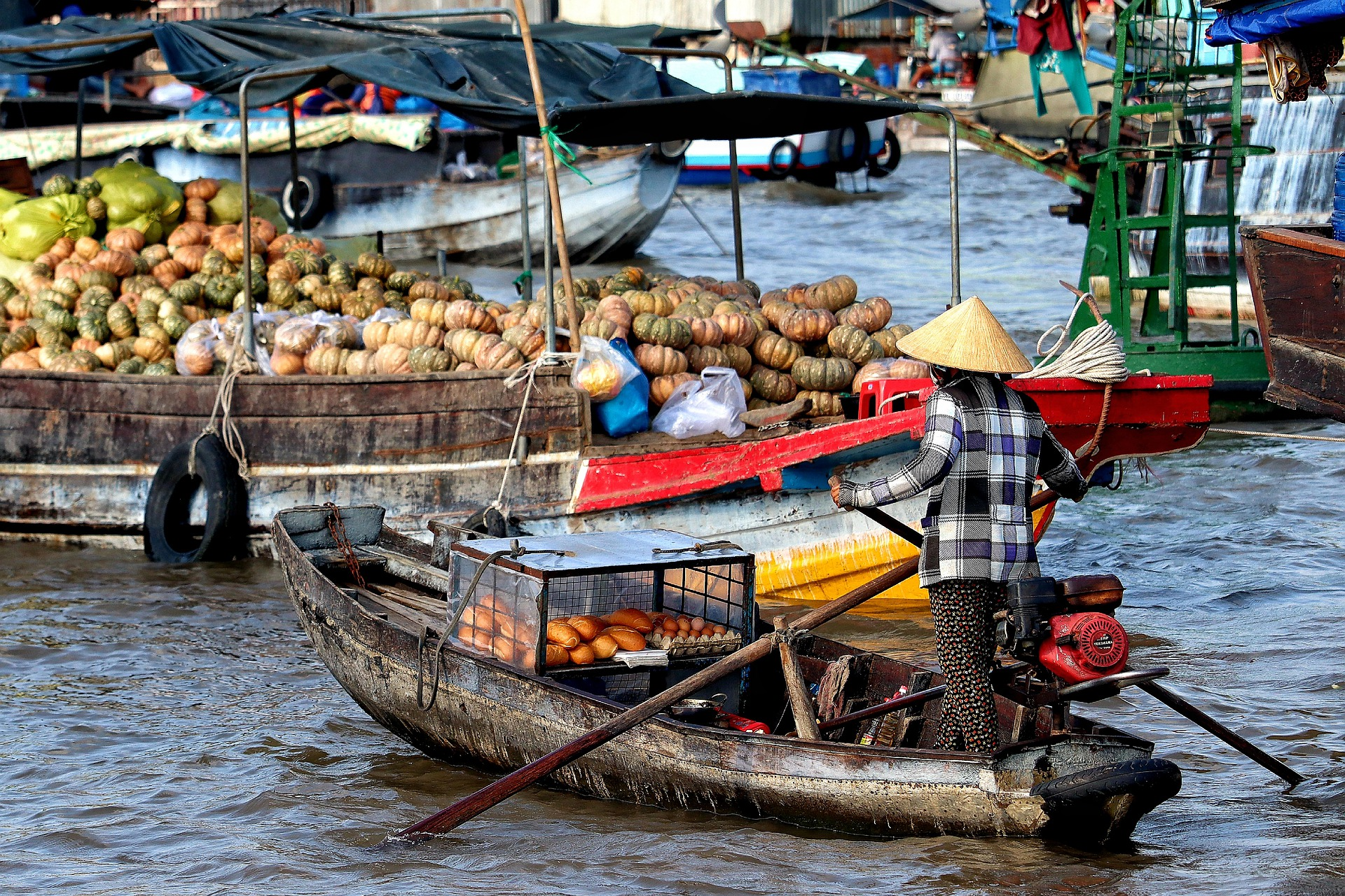 A vendor in a conical hat steers a wooden boat past a large boat filled with pumpkins at a floating market